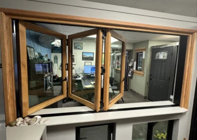 A wooden-framed window opens to an office, showcasing a desk with monitors, walls adorned with pictures, and a brown door. A dog sniffs nearby.
