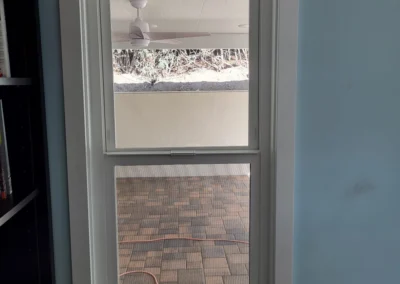 View through a white double-hung window, showing a ceiling fan and a paved outdoor area with an orange extension cord.