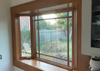 A wooden-framed window in a kitchen, overlooking a garden with a wooden fence and outdoor furniture. Soft natural light fills the space.